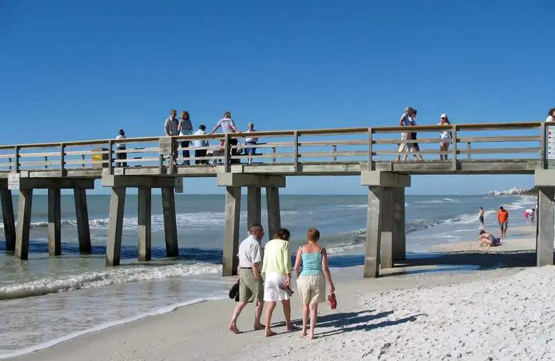 People on the sand beside the Naples Pier under a clear blue sky. The pier deck is closed for a rebuild, so this photo shows an earlier day.