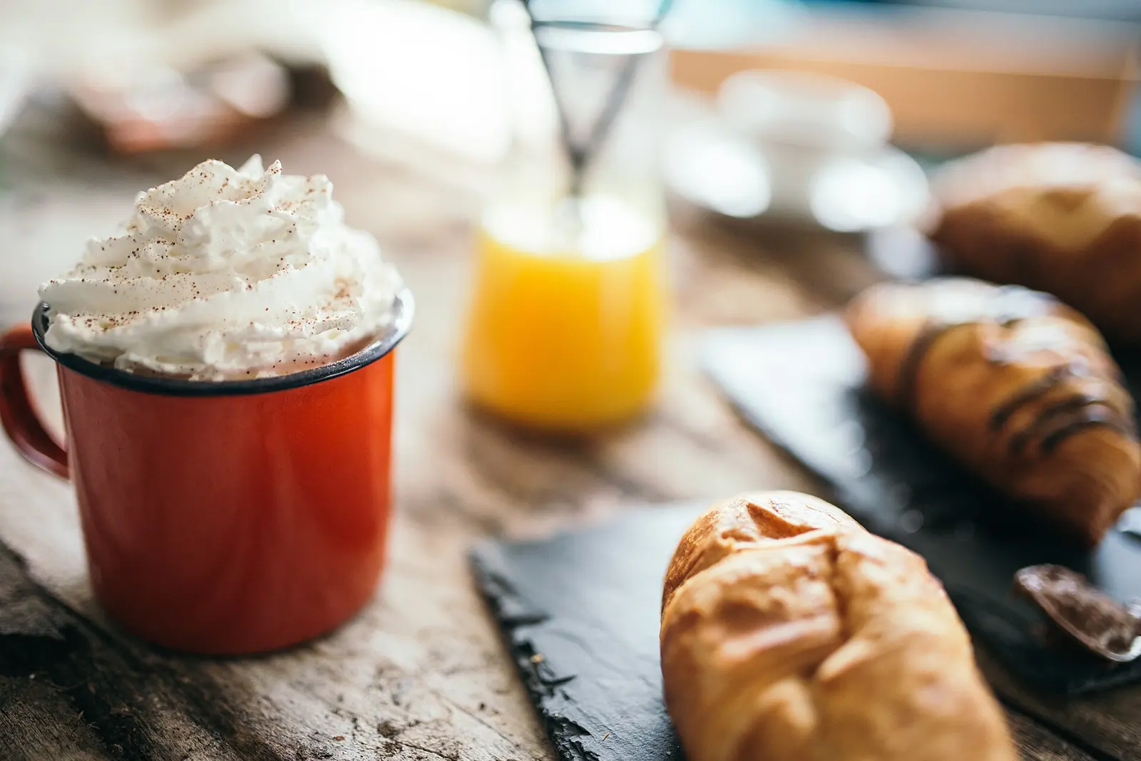 A cozy cafe features a warm red mug filled with whipped cream-topped coffee alongside fresh pastries. A glass of orange juice adds vibrancy to the rustic wooden table.