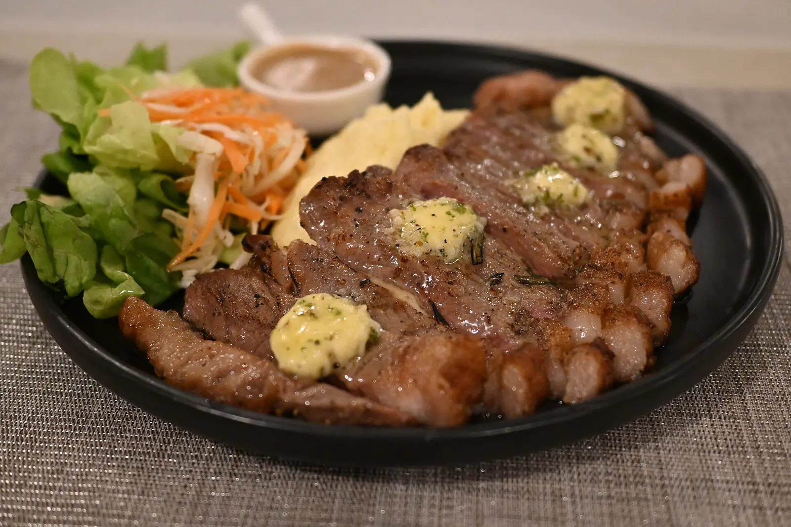 This photo shows a beautifully plated steak dish served on a black plate. The steak appears to be grilled to medium or medium-rare and is topped with small dollops of herb...