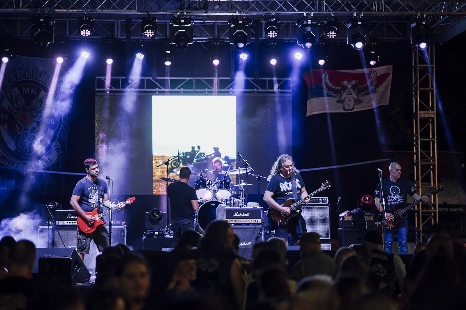 Musicians perform on stage during a lively rock concert at an outdoor venue. The crowd enjoys the music as colorful lights illuminate the scene.